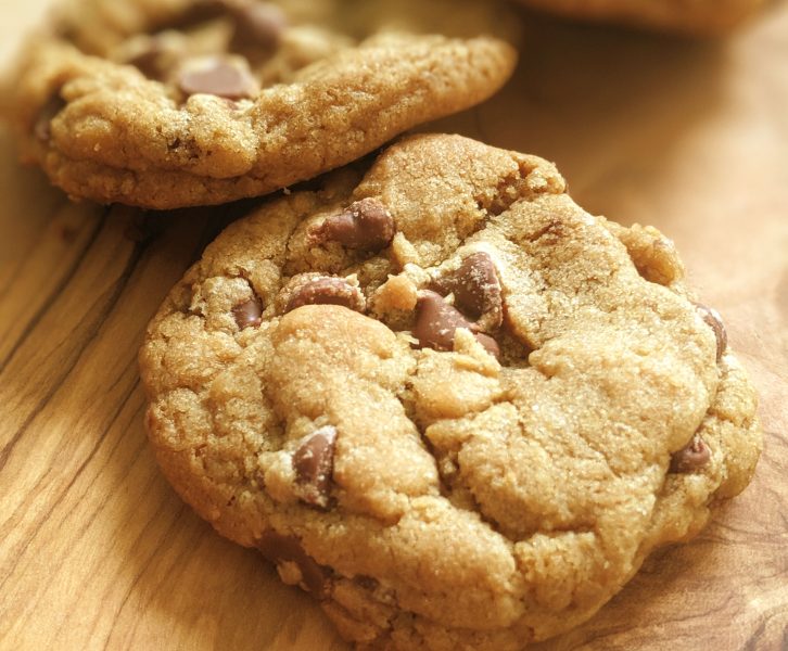 There is a large Cookie with Chocolate Chips on a wooden board with two other cookies in the background